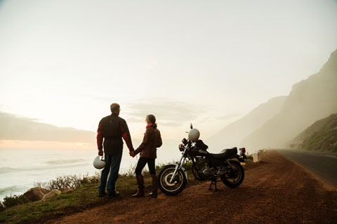 Man and woman stand next to motorcycle and hold hands while watching the sunset.